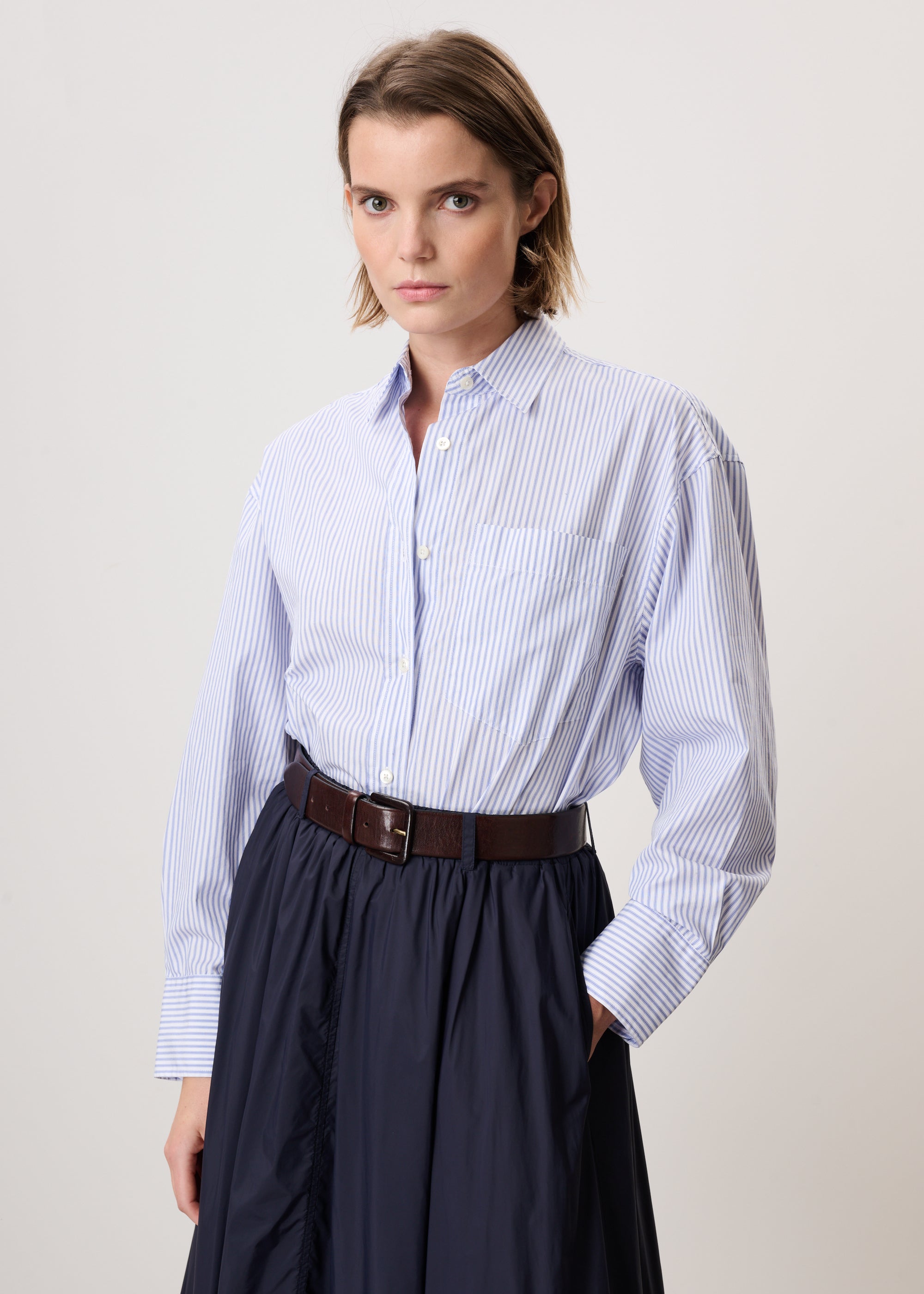 A woman modeling a light blue and white vertically striped button-down shirt tucked into a dark navy skirt, accessorized with a brown belt, against a plain light gray studio background.