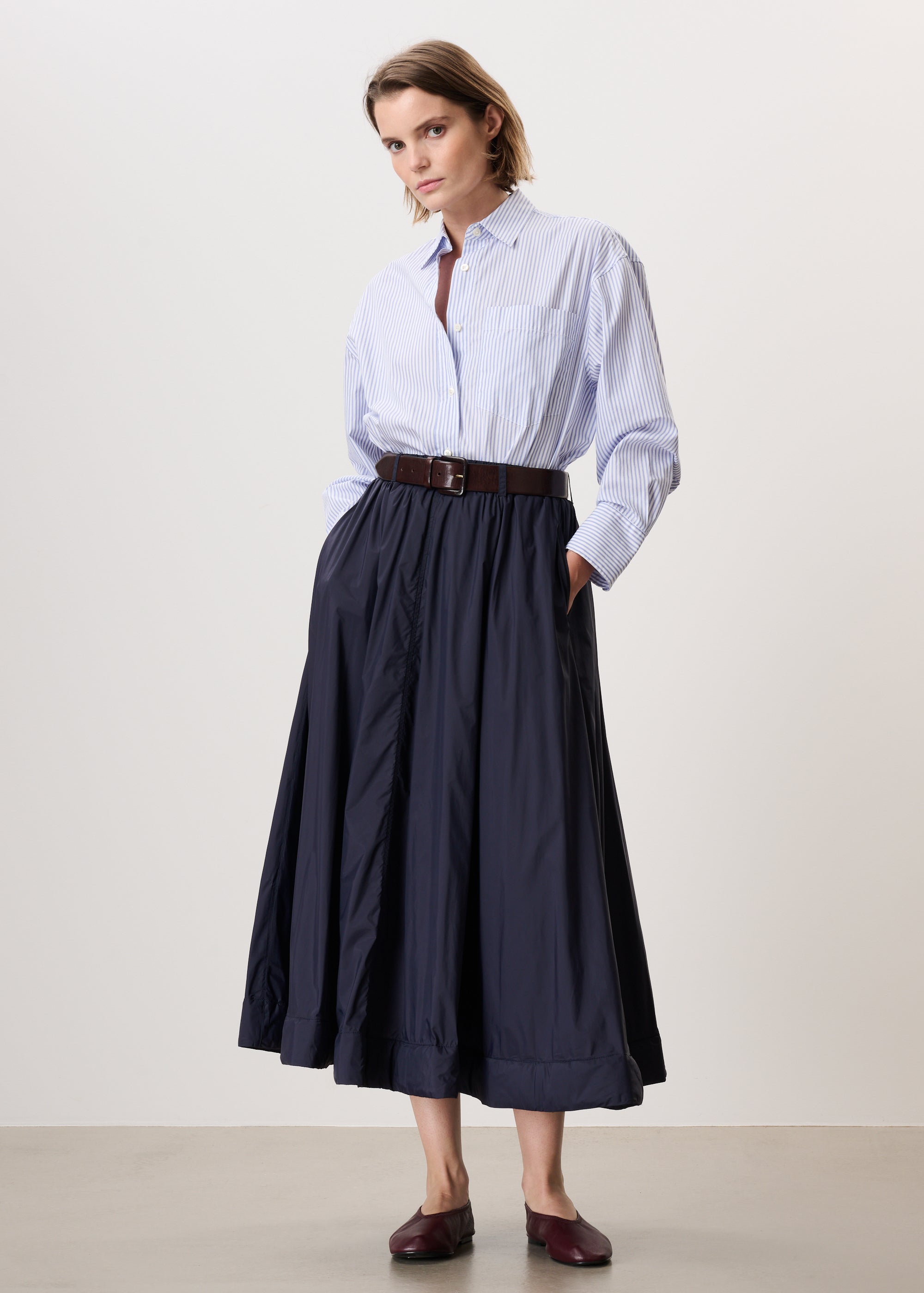 A woman modeling a light blue and white striped button-down shirt paired with a dark navy midi skirt, a brown belt, and dark brown loafers, set against a plain light beige studio background.